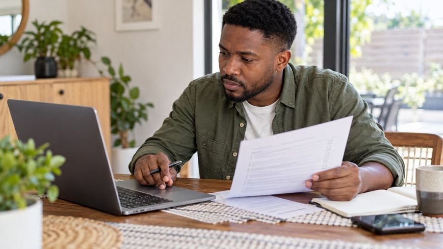 Man checking details on a laptop against a printed checklist — Checking a lender's registration before sharing documents